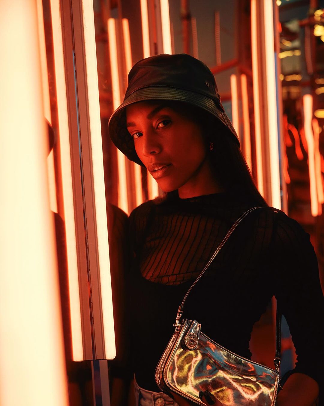 A woman with a bucket hat and crossbody bag poses in front of vibrant orange neon lights, captured in a medium shot.