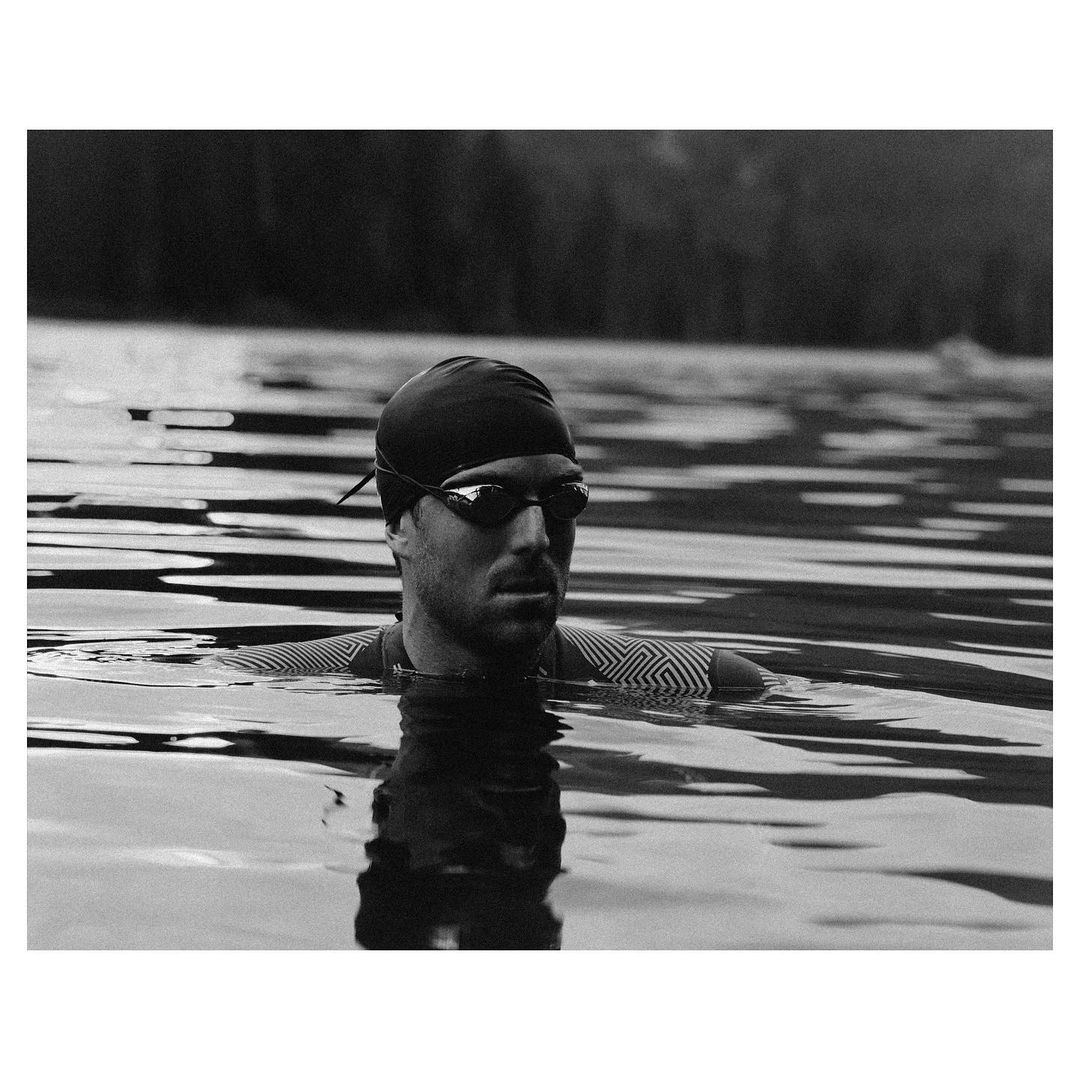 Monochrome close-up of adult male swimmer in lake. He wears goggles and swim cap. Trees are visible in the background.