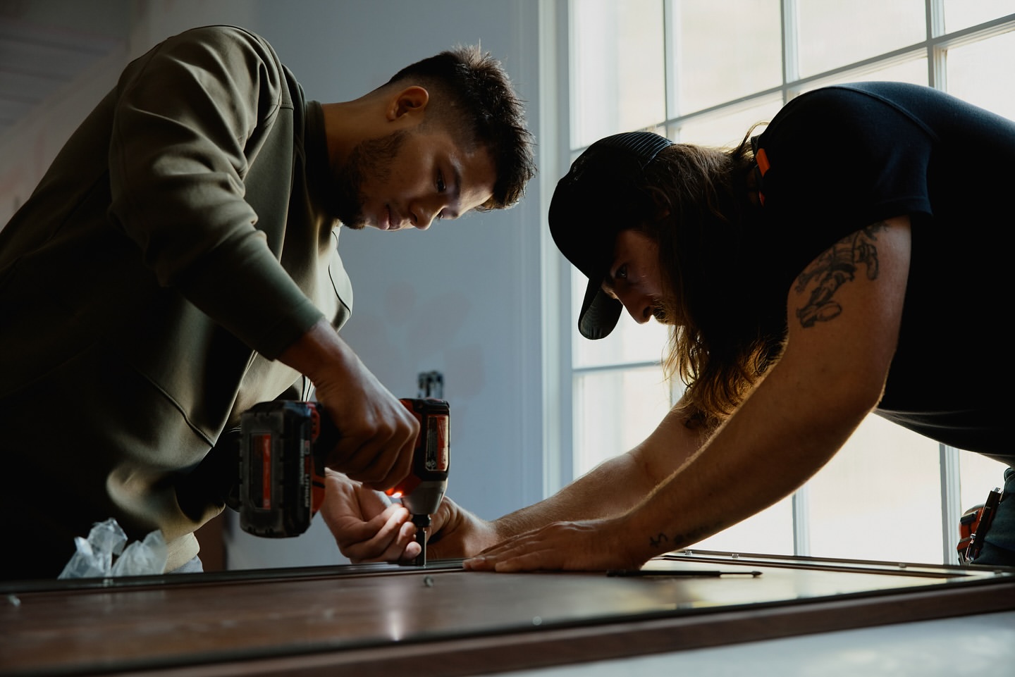 Two adult men in a construction environment use a power drill on a window frame, demonstrating home improvement or carpentry work.