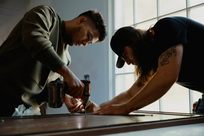 Two adult men in a construction environment use a power drill on a window frame, demonstrating home improvement or carpentry work.