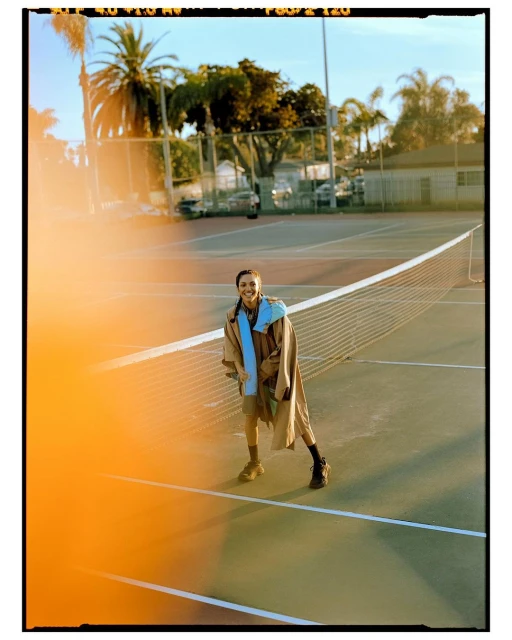 A smiling young woman stands on a tennis court in a tan jacket with palm trees in the background on a sunny day.