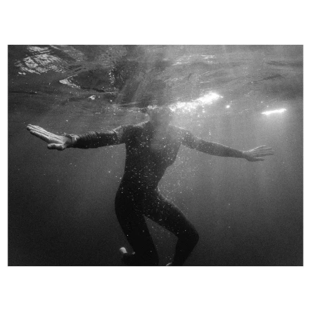 Monochrome underwater photograph of a figure wearing a dark wetsuit floating with arms outstretched beneath the surface in New York