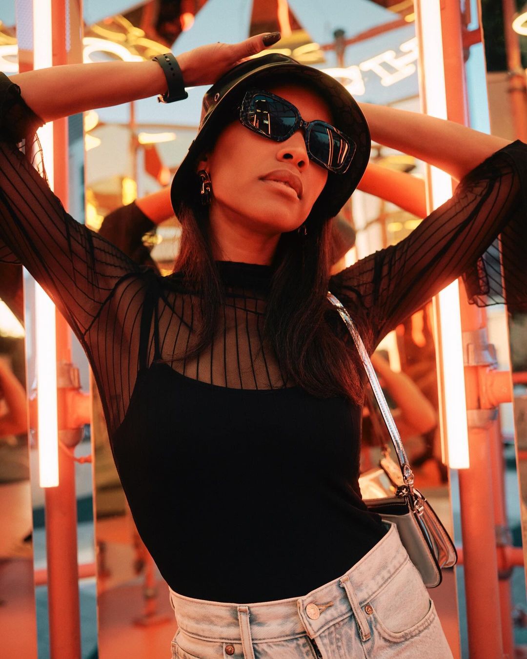 A woman wearing sunglasses and a bucket hat poses in a semi-sheer top and jeans against a blurred backdrop with neon lights.