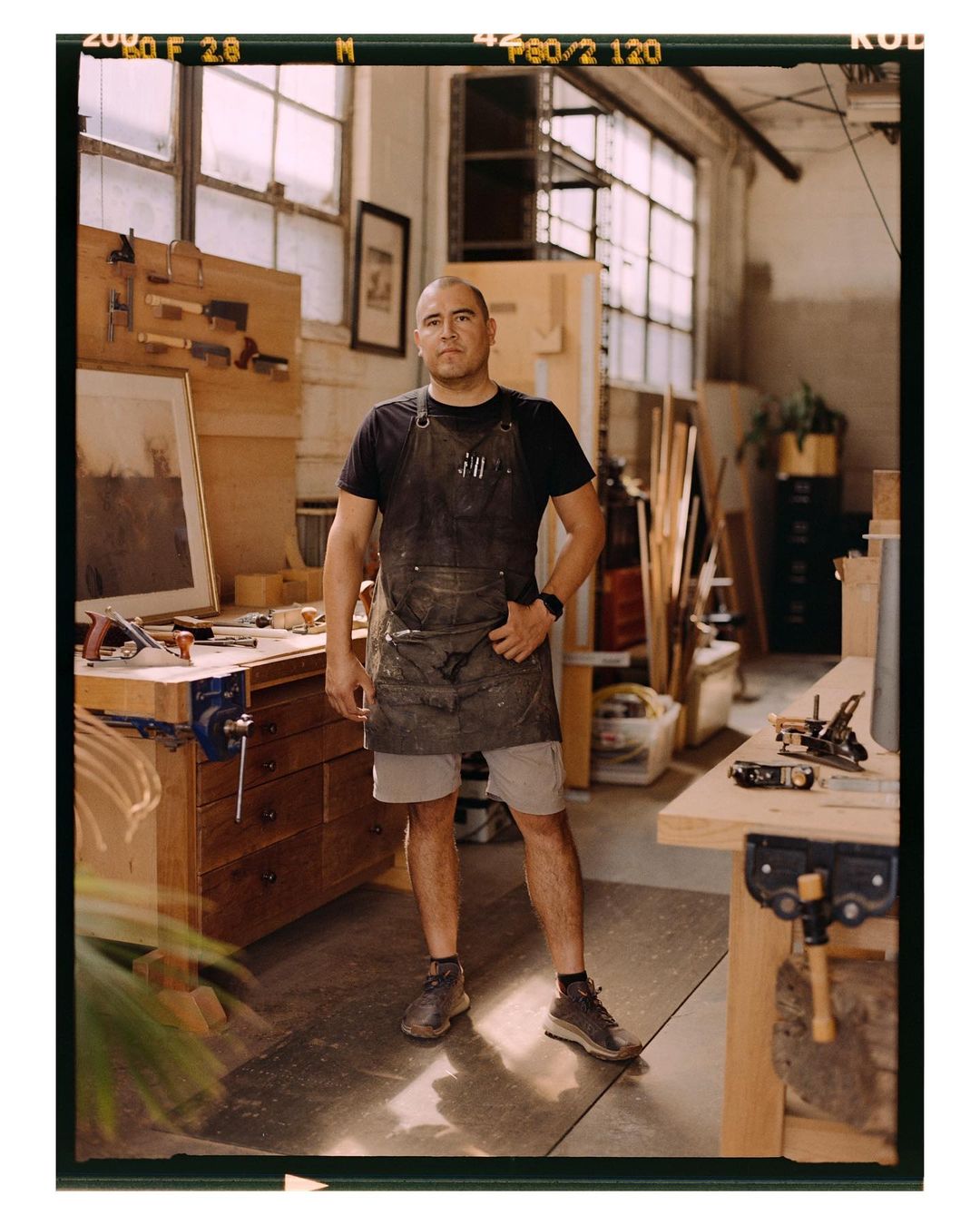 Man in work apron standing in woodworking shop surrounded by tools, wooden surfaces, and natural light from large windows.