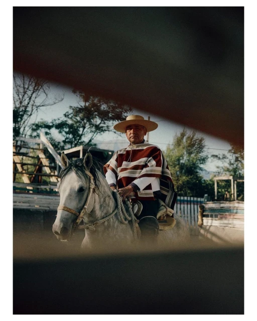 Older adult male riding a grey horse, wearing a hat and poncho, in an outdoor setting with trees, seen through a blurry foreground.