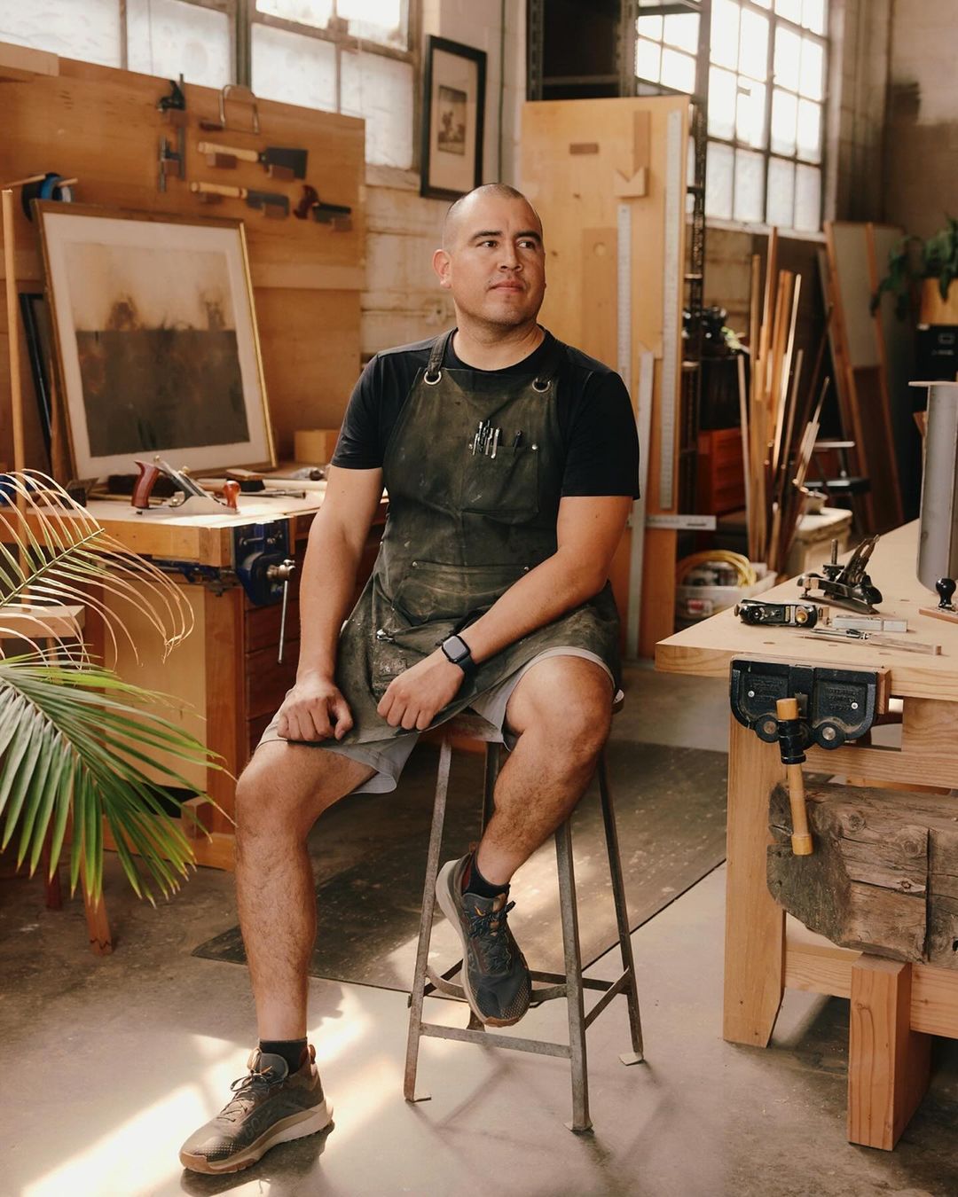 Adult male woodworker wearing an apron sits on a stool in his workshop, surrounded by carpentry tools and wooden work surfaces.