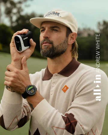 Adult male golfer with beard wearing a beige hat and sweater, using a rangefinder and smartwatch on a golf course on a cloudy day.
