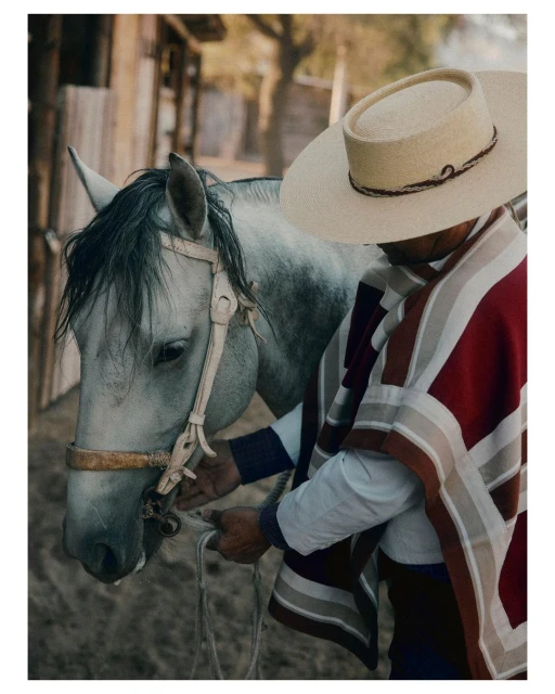 Portrait of a gaucho holding the reins of a grey horse in a rural outdoor setting, wearing a hat and a poncho with red stripes.