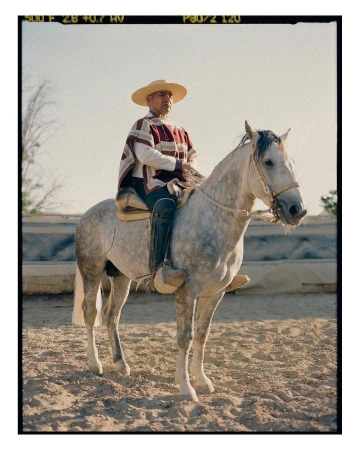 Elderly gaucho in traditional poncho rides a gray horse in sunlit arena, wearing a straw hat and boots, against a clear sky background.