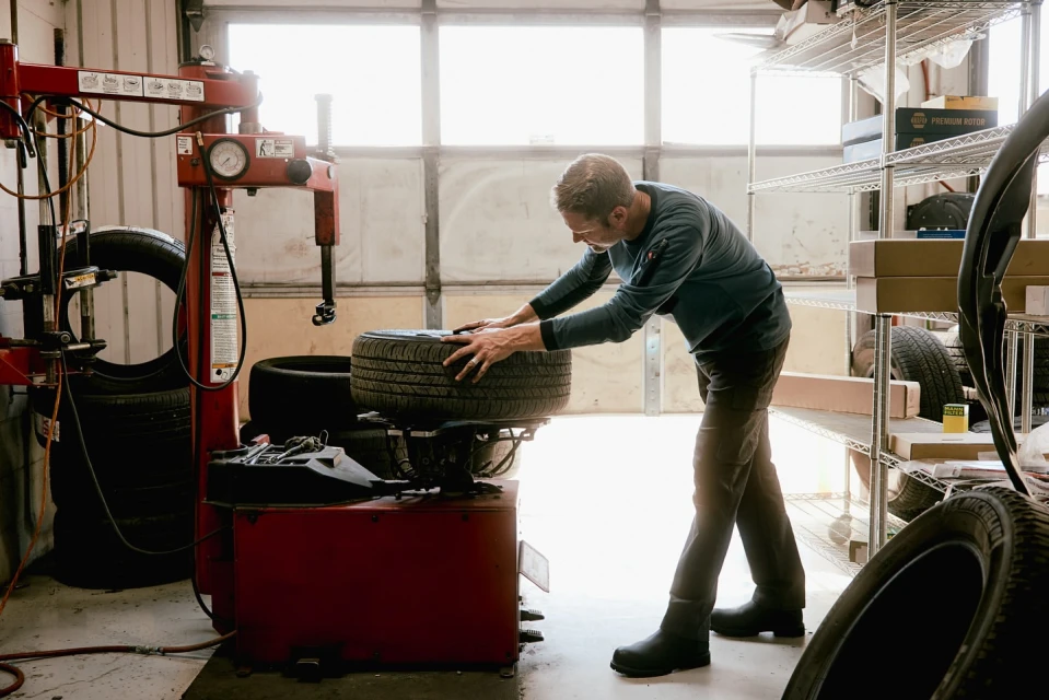 Adult male mechanic working on a tire at a machine in an auto repair shop with tools and tires visible in the background.