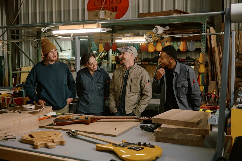Four adults stand in a guitar workshop, smiling and talking. Wood guitar parts, tools, and a 'Guitar Build Workshop' sign are visible.
