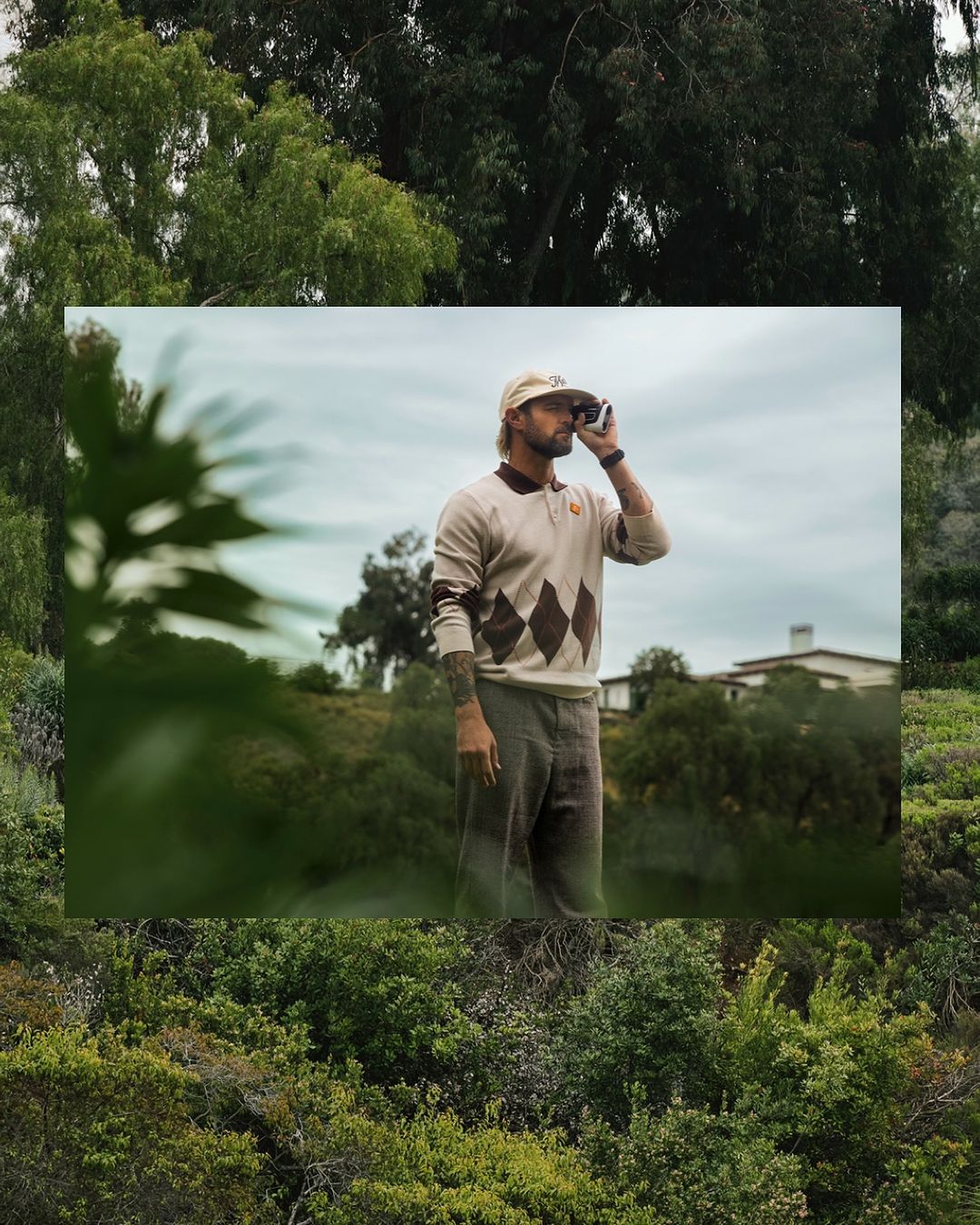 Adult male golfer in argyle sweater holds rangefinder on golf course against natural green background with trees and cloudy sky.