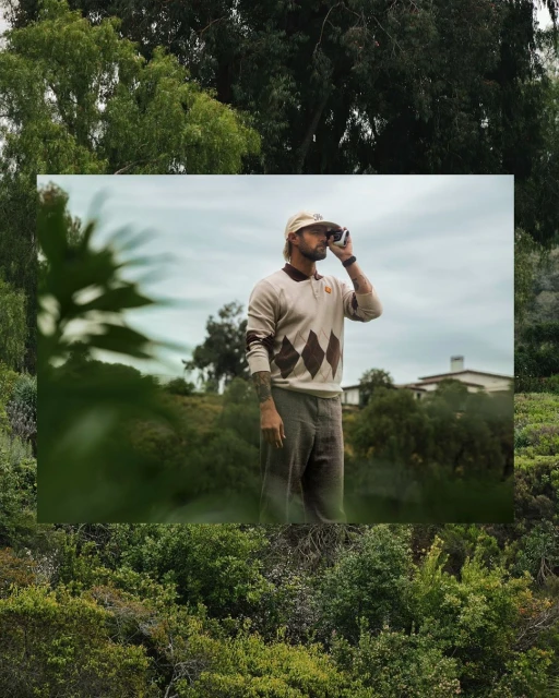 Adult male golfer in argyle sweater holds rangefinder on golf course against natural green background with trees and cloudy sky.