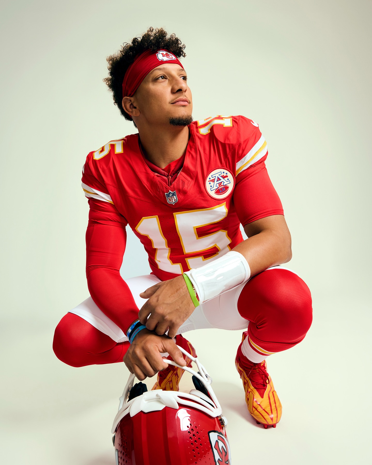 Patrick Mahomes in a red Kansas City Chiefs uniform poses with a helmet, looking upward, in a studio shot with soft lighting.