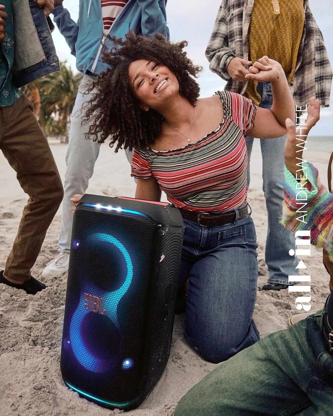 Young woman smiles and dances on a beach with friends, holding a portable JBL speaker with illuminated rings, under a bright sky.