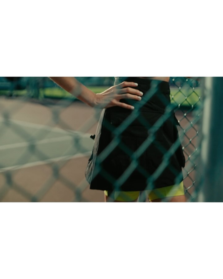 A partial view of an adult woman on a tennis court is seen through a chain-link fence. She wears a black skirt and yellow shorts.