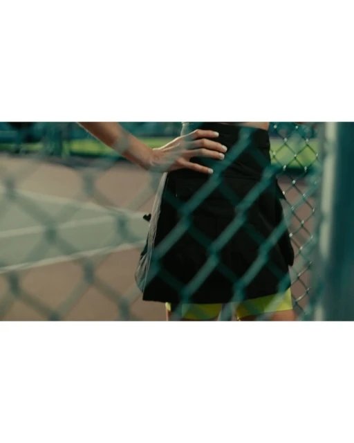 A partial view of an adult woman on a tennis court is seen through a chain-link fence. She wears a black skirt and yellow shorts.