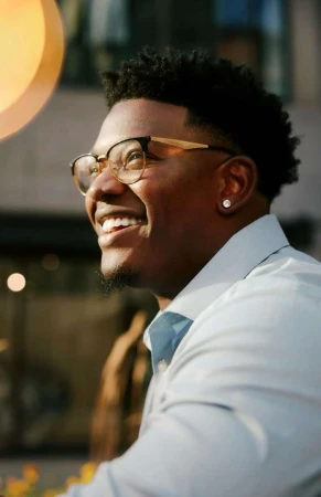 Portrait of a smiling Black man wearing glasses and a light button-down shirt outdoors in New York.