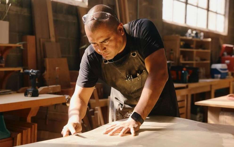 A man works with wood in a workshop, wearing safety glasses and an apron. The scene is illuminated with natural light from a window.