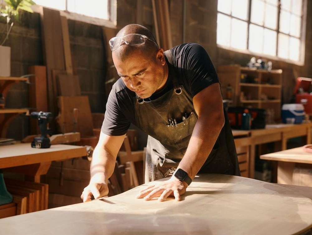 A craftsman is smoothing a wooden plank in a workshop illuminated by natural light, wearing safety glasses and a leather apron.