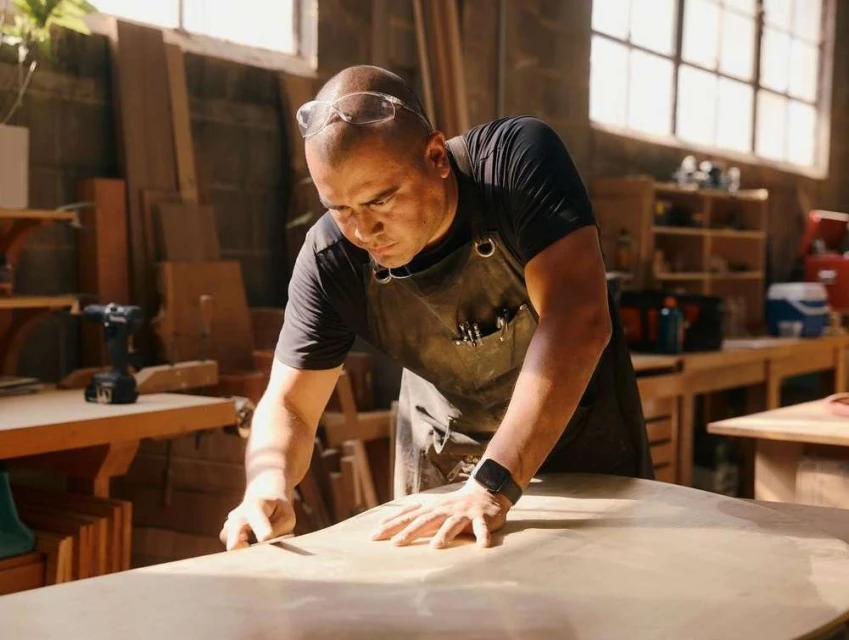 A craftsman is smoothing a wooden plank in a workshop illuminated by natural light, wearing safety glasses and a leather apron.