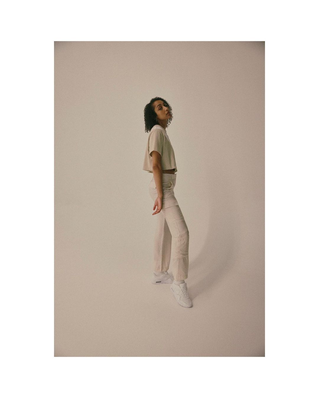 Studio shot of a young adult female model in a beige crop top and off-white pants posing in front of a neutral backdrop.