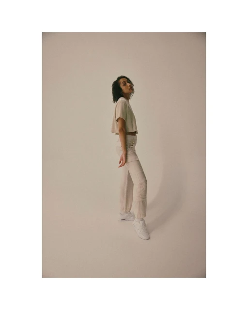 Studio shot of a young adult female model in a beige crop top and off-white pants posing in front of a neutral backdrop.