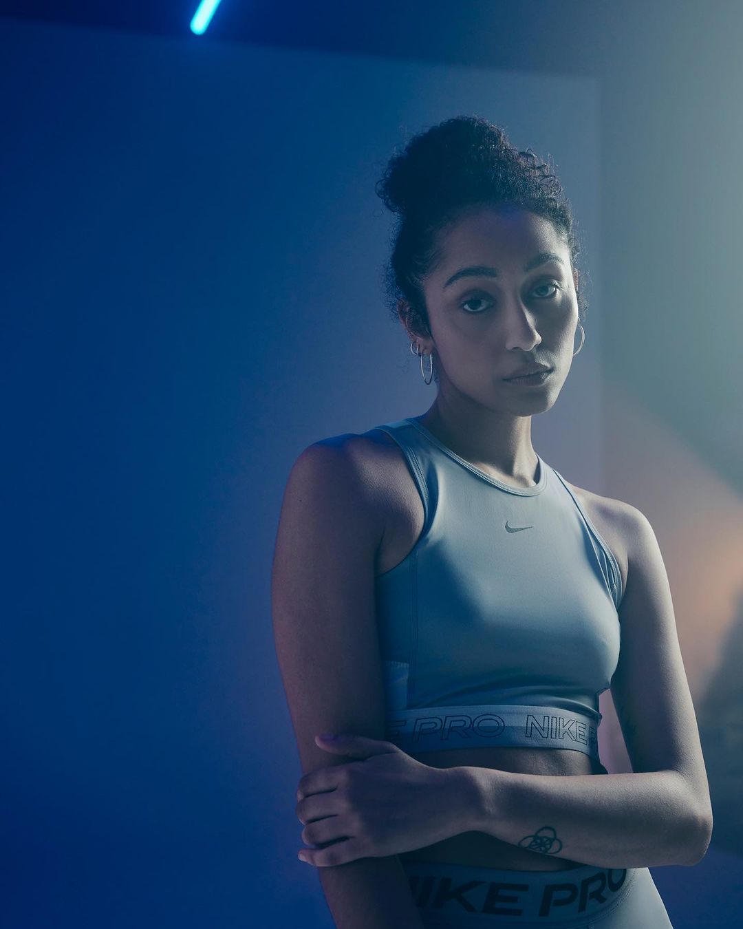 Portrait of a woman with her hair in a bun, wearing a sports bra and athletic pants with Nike Pro branding, against a blue background.