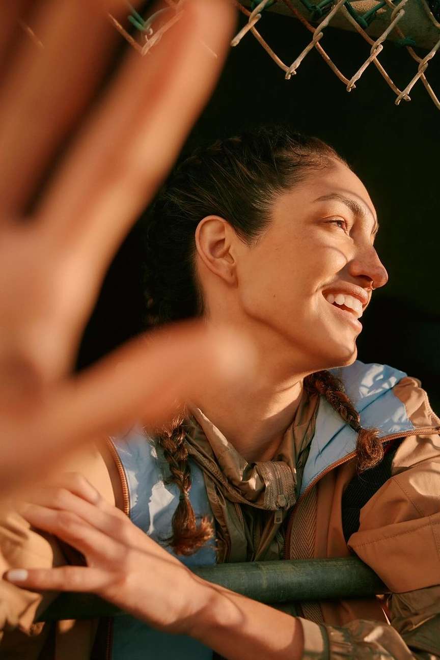 A smiling woman with braids in outdoor clothing is captured in a close-up shot. Her hand is raised, partially obscuring her face.