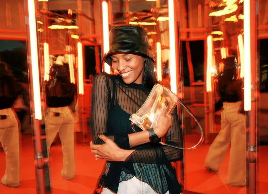 Smiling woman in a black bucket hat holds a transparent handbag, standing in front of mirrors with orange neon lights reflecting around her.
