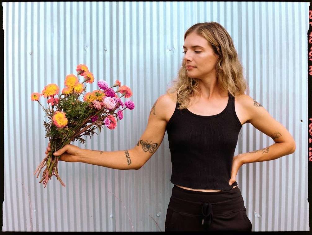Blonde woman holds a colorful bouquet of flowers. She wears a black tank top and stands in front of a blue corrugated metal wall.
