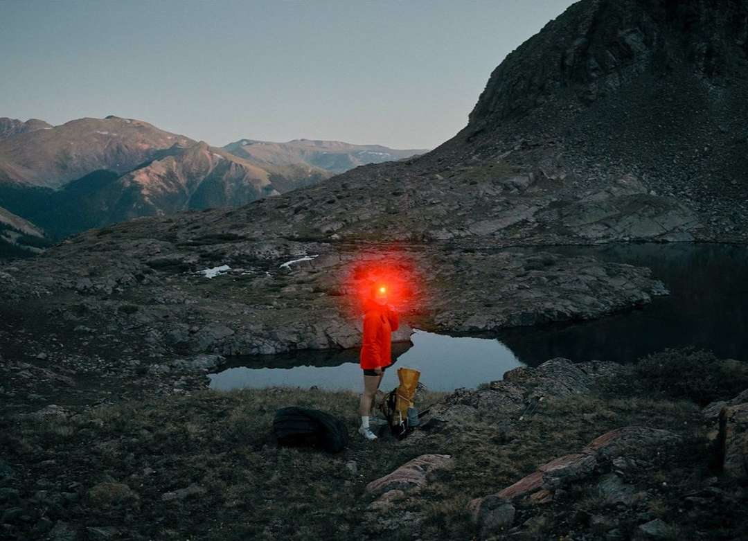 Person with red headlamp hiking in rugged mountain terrain near a lake at dusk, carrying a backpack and accompanied by a dog.