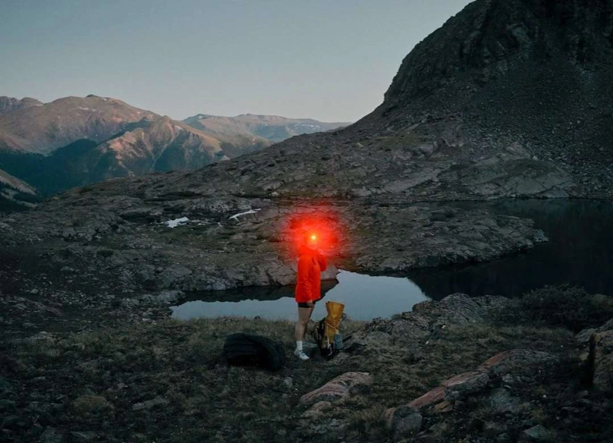 Person with red headlamp hiking in rugged mountain terrain near a lake at dusk, carrying a backpack and accompanied by a dog.
