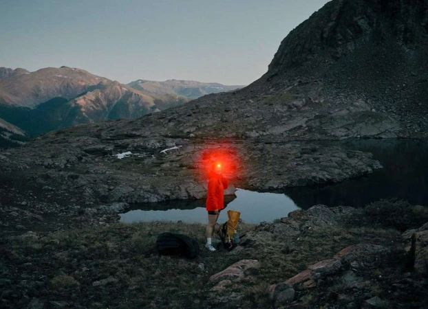 Person with red headlamp hiking in rugged mountain terrain near a lake at dusk, carrying a backpack and accompanied by a dog.