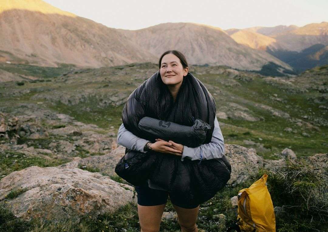 Woman in sleeping bag stands in mountainous landscape smiling, wearing shorts and jacket.  Sunlit mountain range in the distance.