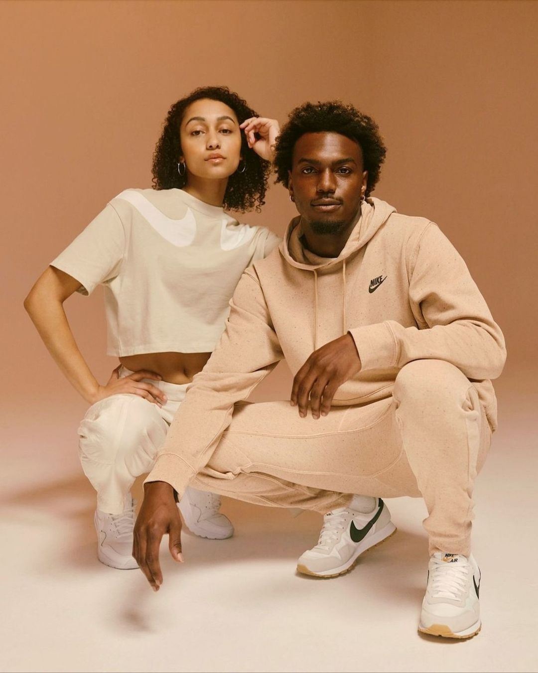 Two models posing in matching beige sportswear and white sneakers against a warm neutral studio backdrop.