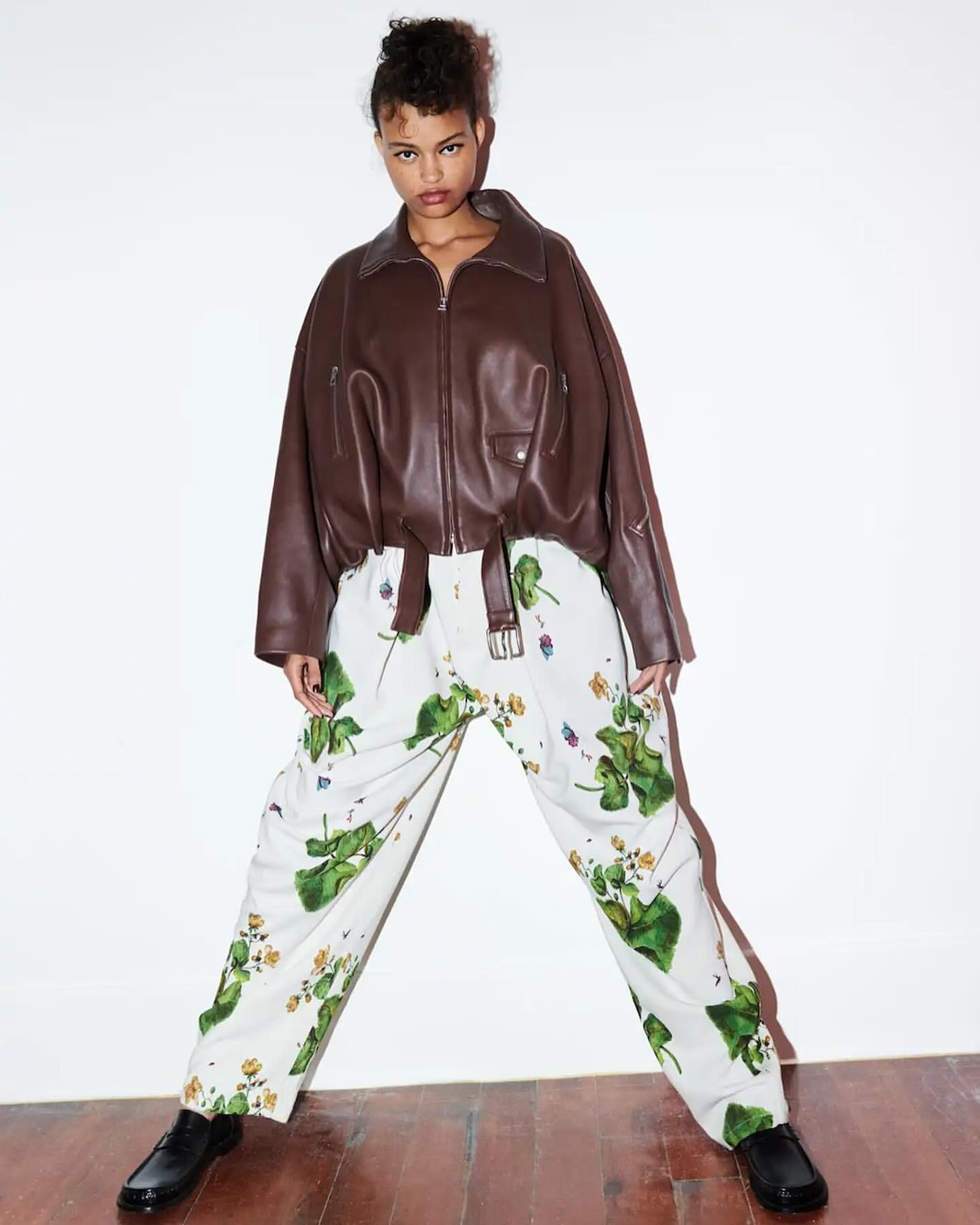 A model poses in a brown leather jacket and floral pants in a bright studio, offering a modern, edgy fashion statement.