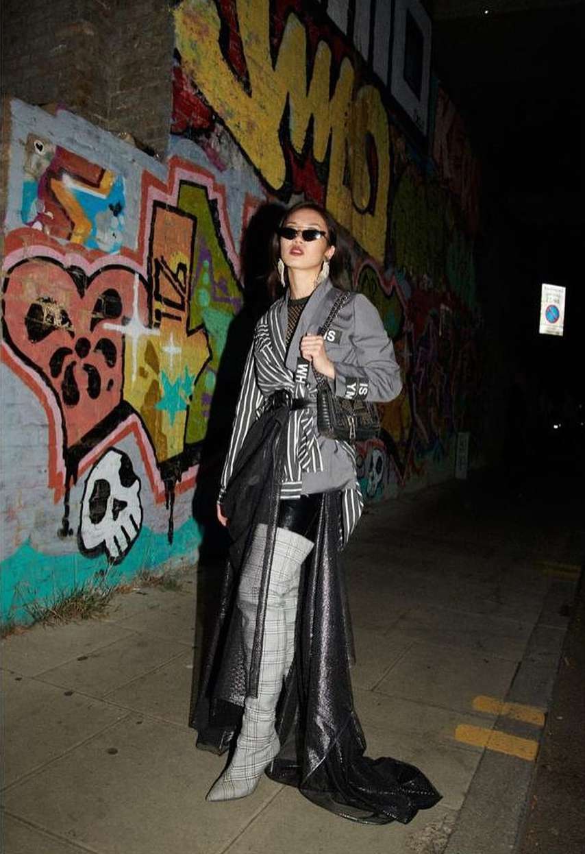 Woman poses in trendy street style against graffiti backdrop in city night scene, wearing a blazer, skirt, thigh-high boots, and sunglasses.