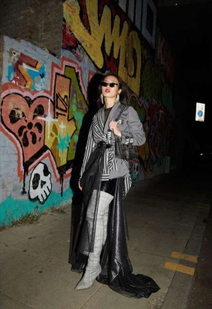 Woman poses in trendy street style against graffiti backdrop in city night scene, wearing a blazer, skirt, thigh-high boots, and sunglasses.