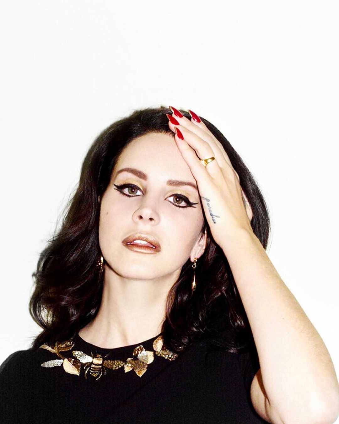 Close-up portrait of a dark-haired woman with red nails and a statement necklace, posing against a white background in a studio setting.