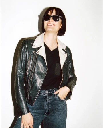 Woman with dark bob and sunglasses wearing a dark leather jacket and denim jeans against a plain white background.
