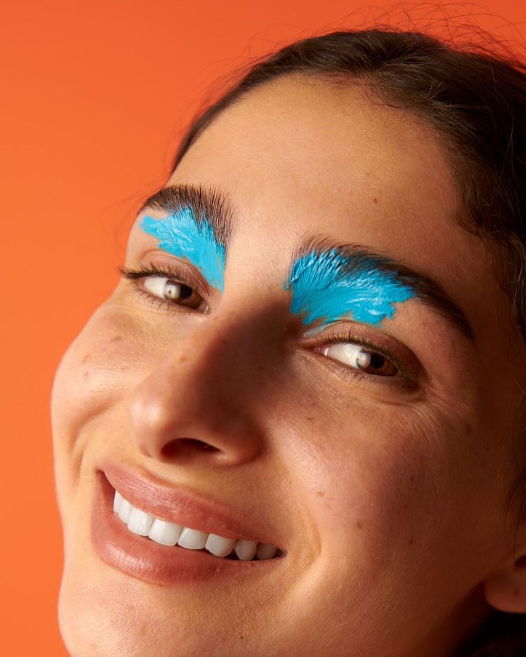 Close-up portrait of a smiling adult woman with bright turquoise paint on her eyebrows, set against a solid orange background.