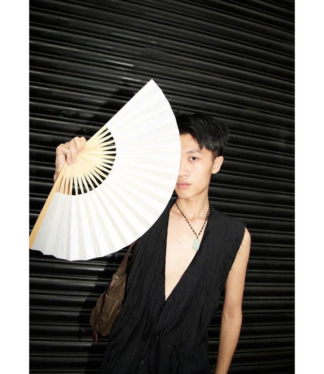 Young adult poses against a dark background with an open white hand fan and wearing a black sleeveless shirt with a brown shoulder bag.