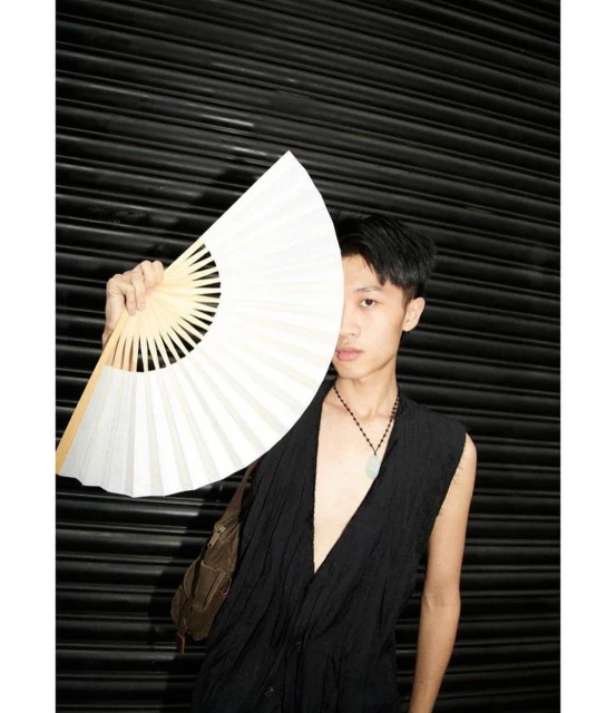 Young adult poses against a dark background with an open white hand fan and wearing a black sleeveless shirt with a brown shoulder bag.