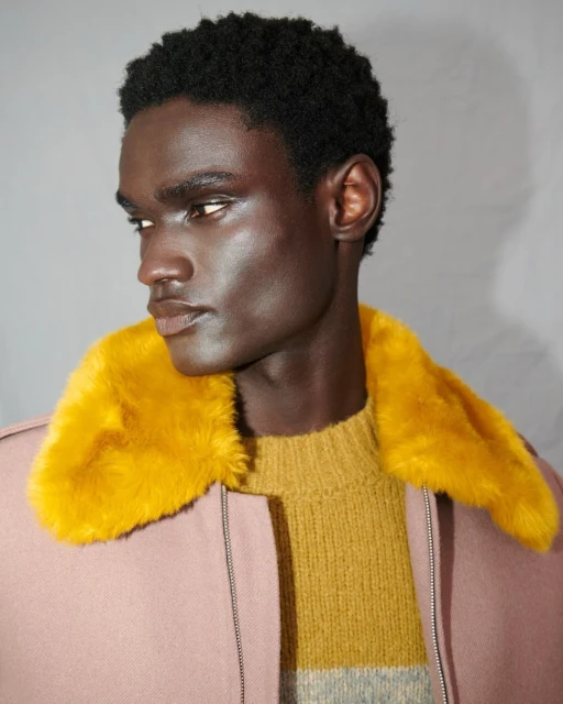 Studio portrait of a Black male model in a pink jacket with a yellow faux fur collar and mustard yellow knit sweater, looking to the side.