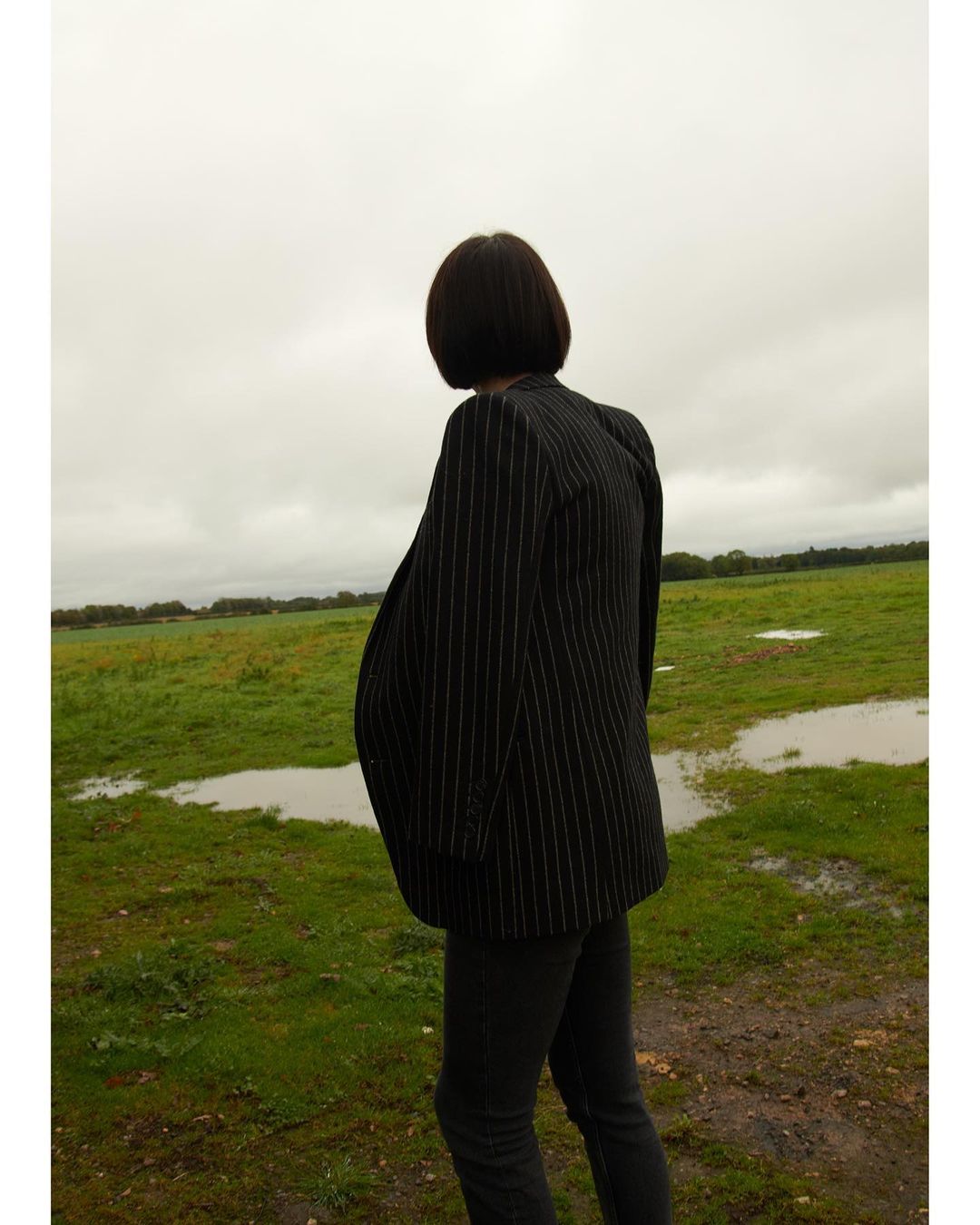 An adult stands in a field with puddles under a cloudy sky. The person wears a striped blazer and dark trousers in a rural setting.