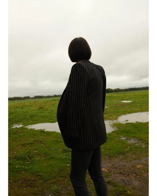 An adult stands in a field with puddles under a cloudy sky. The person wears a striped blazer and dark trousers in a rural setting.