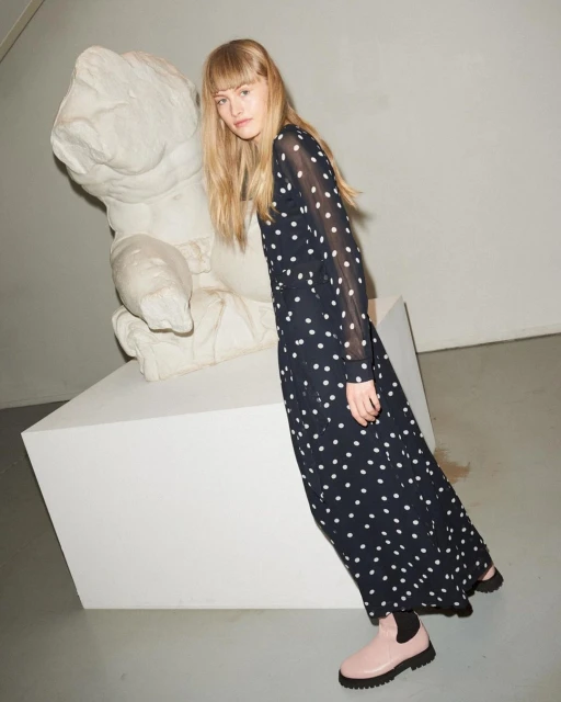 A blonde woman in a polka dot dress poses indoors with a white sculpture. She's leaning on a cubic stand, wearing shiny pink boots.