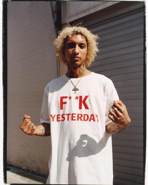 Adult male with fair skin, curly blonde hair, and tattoos wearing a white tee shirt with a red text graphic stands outside.
