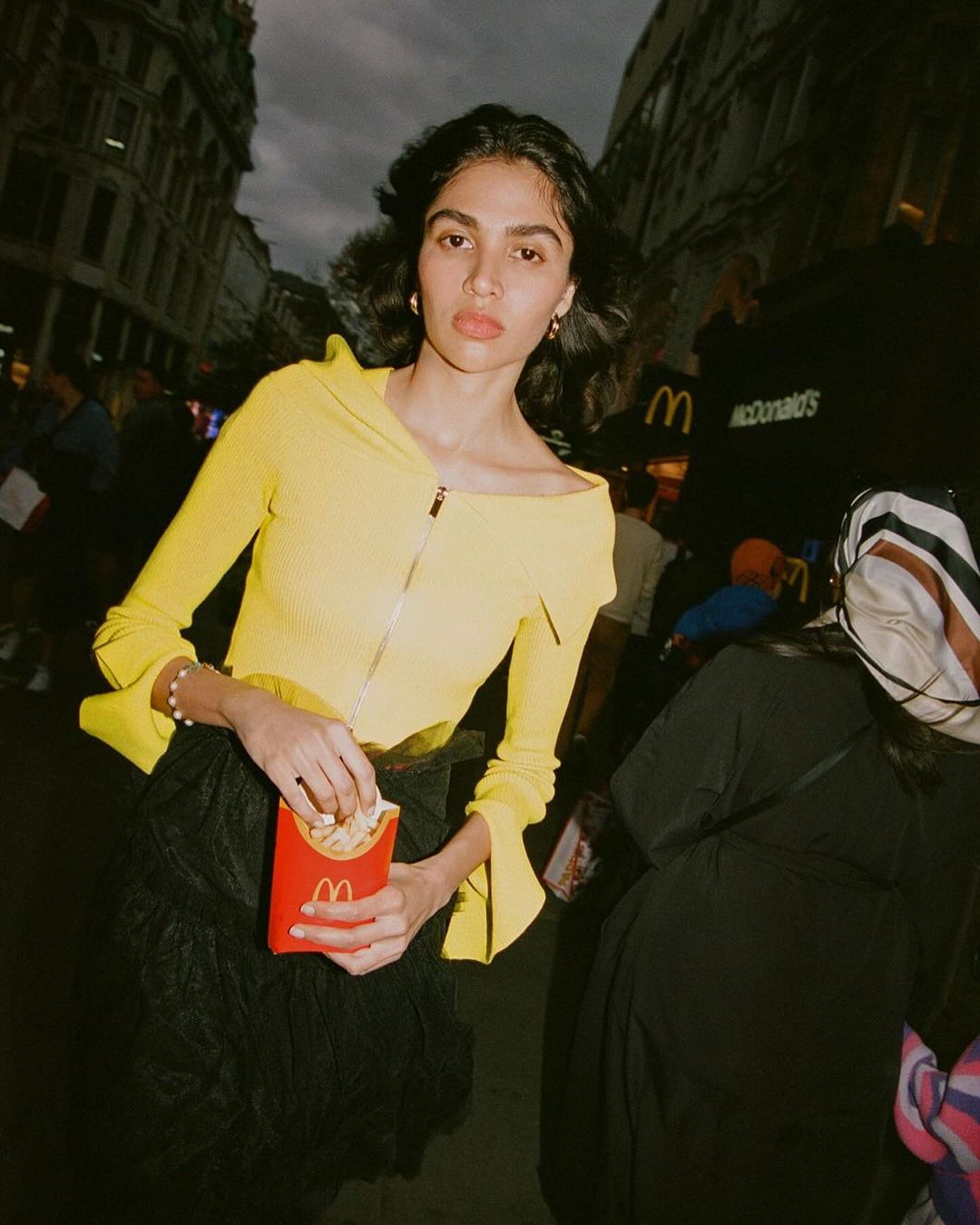 A young woman in a yellow top eats French fries in a city street scene, with a McDonald's restaurant sign visible in the background.